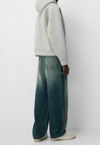 Ljusgrå oversized hoodie som bärs med vida blå denimjeans med en bleknande effekt och vita sneakers. Bakifrån.