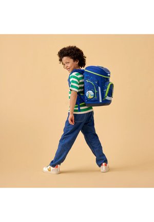 Young child wearing blue and green striped shirt, blue pants, white sneakers, walking with a large blue backpack decorated with patterns and a badge.