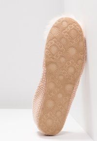 Růžová pletená domácí obuv s vzorovanou béžovou semišovou podrážkou. Obsahuje huňatý lem podél okraje, který přidává texturu a detail.