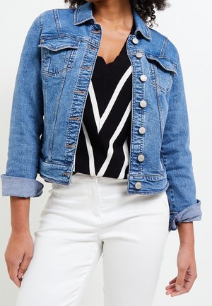 Mujer que lleva una chaqueta vaquera azul claro remangada sobre un top con patrón en blanco y negro, combinada con pantalones blancos de talle alto.