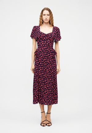 Donna con capelli mossi che indossa un vestito midi blu navy con motivo floreale rosso, maniche corte svolazzanti e tacchi neri con cinturini, in piedi su sfondo bianco.