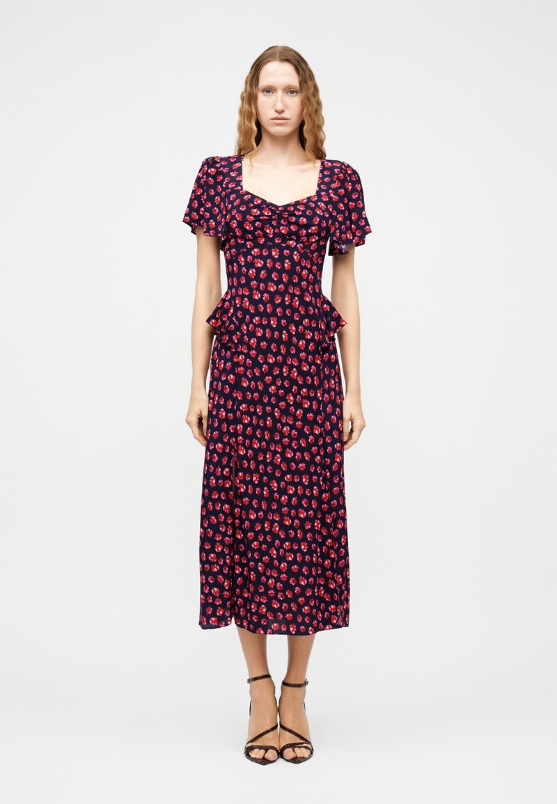 Γυναίκα με κυματιστά μαλλιά που φοράει ένα μπλε σκούρο midi φόρεμα με κόκκινο ανθισμένο μοτίβο, κοντά φουσκωτά μανίκια και μαύρες πέδιλες με λουράκια, στέκεται μπροστά σε λευκό φόντο.