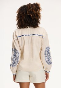 Blusa bianca sporca con dettagli ricamati blu sulle maniche e sul retro, caratterizzata da un tessuto testurizzato e una vestibilità comoda, abbinata a pantaloni corti chiari.