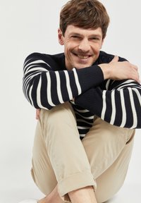 Maglione a righe navy e bianche in maglia con bottoni sulla spalla, indossato con pantaloni beige arrotolati e scarpe bianche, creando un look casual.