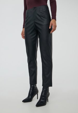 Donna in pantaloni neri in pelle a vita alta e stivaletti neri con punta a stiletto su uno sfondo neutro.