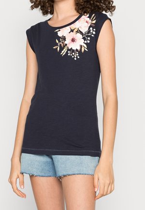 Kobieta w granatowym bezrękawniku z różowym kwiatowym nadrukiem przy dekolcie i jasnoniebieskich szortach jeansowych z postrzępionymi brzegami.