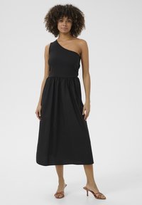 Svart klänning med en axel i ribbat tyg, med en åtsittande överdel och en svävande kjol. Sandaler med remmar framhäver looken.