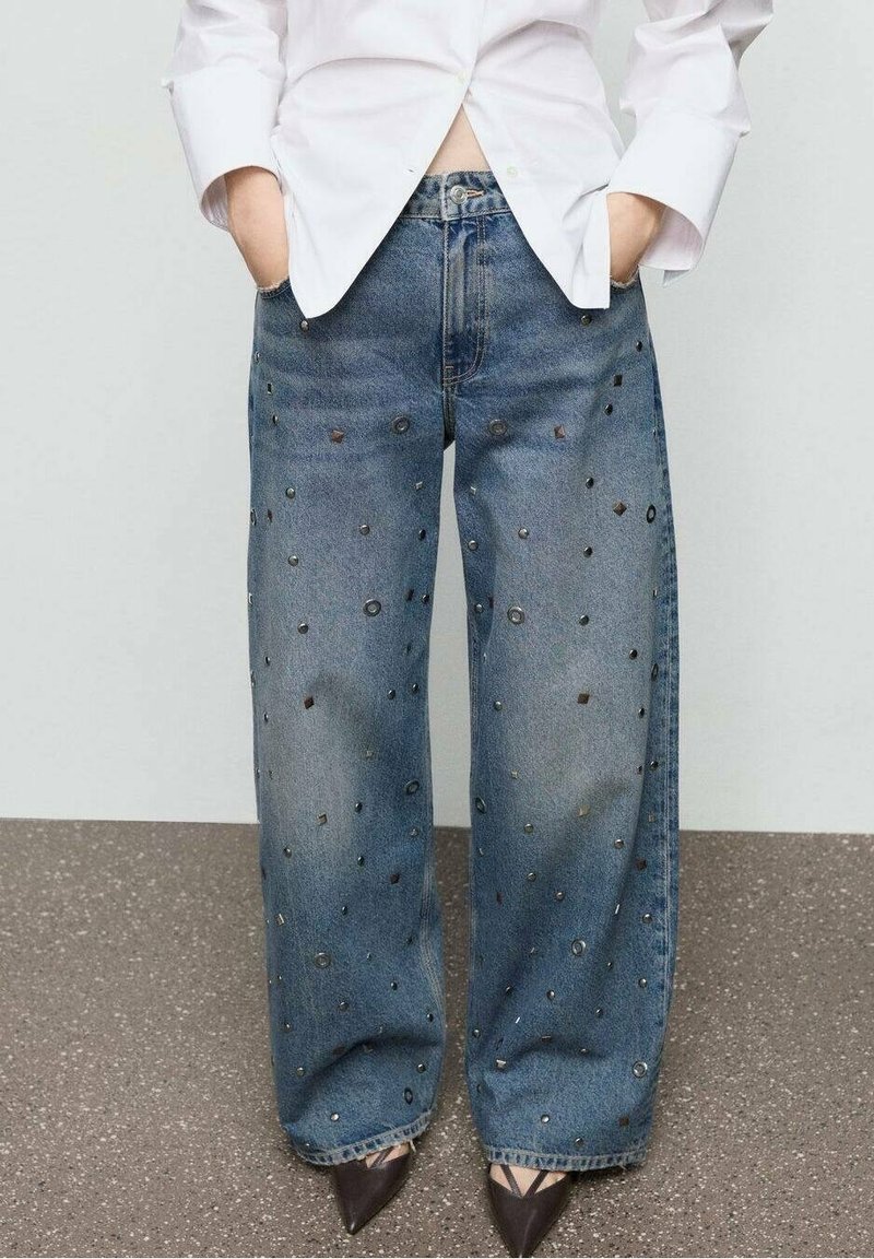 Jean bleu à jambes larges avec des petits clous métalliques éparpillés, associé à une chemise blanche boutonnée et des talons noirs pointus.