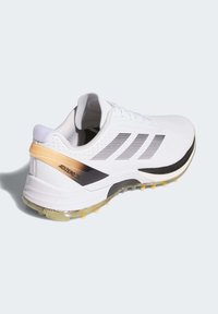 Valkoinen urheilukenkä mustilla ja harmailla raidoilla, teksturoidulla pinnalla, kultaisilla korostuksilla ja tukevalla pohjalla, jossa näkyvät pito-ominaisuudet.