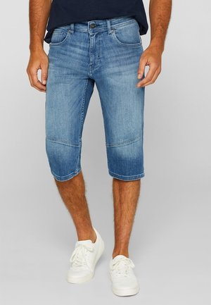 Mann trägt knielange blaue Jeansshorts, weiße Turnschuhe und ein dunkelblaues Hemd, steht vor einem schlichten grauen Hintergrund.