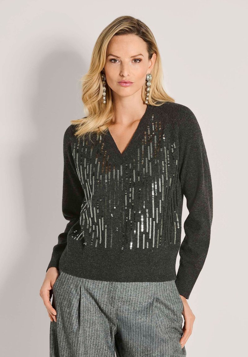 Frau mit langen blonden Haaren, die einen dunkelgrauen, mit Pailletten besetzten V-Ausschnitt-Pullover und graue Streifenanzughosen trägt, steht vor einem schlichten hellen Hintergrund.