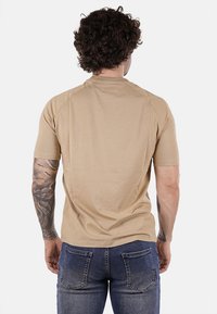 Maglietta beige a maniche corte in cotone, con collo rotondo e una texture liscia. Tatuaggi visibili sulle braccia. Indossata con jeans in denim.