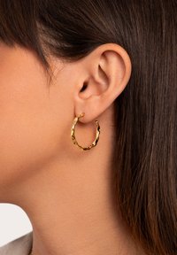 Pendiente de aro de oro con un diseño retorcido, que cuenta con una superficie texturizada y un tamaño mediano, exhibido en la oreja de un modelo con cabello oscuro.