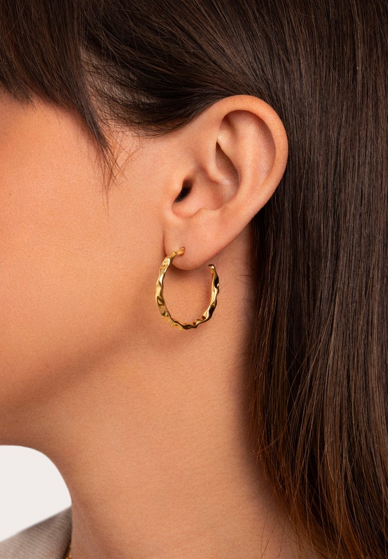 Pendiente de aro de oro con un diseño retorcido, que cuenta con una superficie texturizada y un tamaño mediano, exhibido en la oreja de un modelo con cabello oscuro.