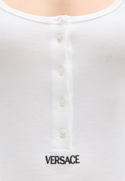 Κοντινό πλάνο λευκού ριμπ ύφανσης τοπ με πέντε κουμπιά στο κέντρο και μαύρο κεντητό λογότυπο "VERSACE" κάτω από τα κουμπιά.