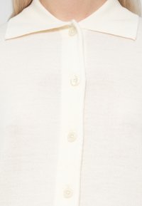 Primer plano de un cárdigan de punto en crema pálido con pequeños botones redondos y un cuello ancho en una persona.