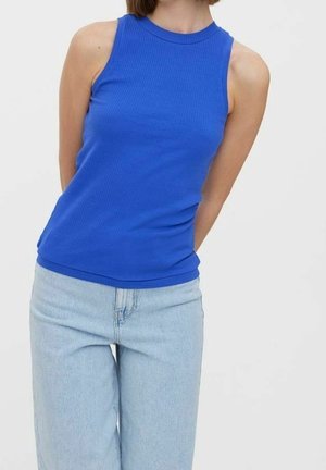 Vrouw draagt een mouwloze koningsblauwe geribbelde top en lichtblauwe hoog getailleerde spijkerbroek, staand tegen een effen lichte achtergrond.