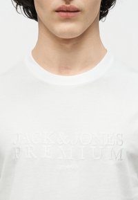 Νεαρό άτομο που φοράει λευκό μπλουζάκι με στρογγυλή λαιμόκοψη και ανάγλυφο λευκό κείμενο στο στήθος που γράφει "JACK & JONES PREMIUM EST. 1990".