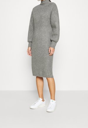 Persona que lleva un vestido midi de punto gris acanalado de manga larga y zapatillas blancas, de pie contra un fondo liso y claro.