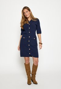 Marineblå kjole med korte ærmer, knapper foran, to brystlommer og en tætsiddende pasform. Kombineret med lysebrune ruskindsstøvler op til knæene.