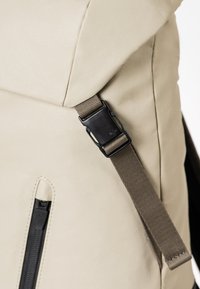 Beige Rucksack aus strukturiertem Material, mit umklappbarem Deckel, schwarzem Metallzubehör und einem braunen verstellbaren Trageriemen. Verfügt über eine Reißverschlusstasche.
