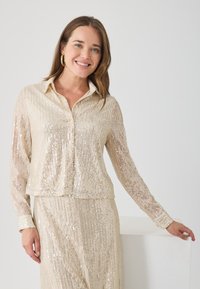 Bluză din mătase bej cu mărgele, cu mâneci lungi, design cu guler și model cu dungi verticale. Textură netedă cu un efect subtil de strălucire.