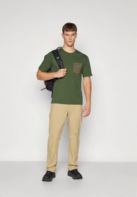 Žalia trumpomis rankovėmis marškinėliai su chaki spalvos krūtinės kišene, derinami su smėlio spalvos kelnėmis ir juodais batais. Asmuo nešioja juodą kuprinę.