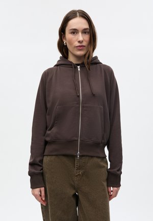 Jonge vrouw met een bruine hoodie met rits en olijfgroene broek, staande tegen een effen lichte achtergrond.