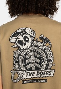 Beige tričko s černým grafikou lebky v čepici, která drží nářadí a dřevěné prkna, s textem „THE DOERS“ níže.