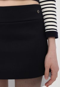 Zwarte mini-rok met een gladde stoftextuur, voorzien van een decoratieve metalen bloemdetail bij de tailleband en een doorschijnende visnetlaag aan de zoom.