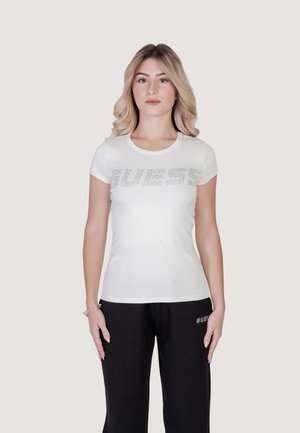 T-shirt en coton blanc à manches courtes, arborant un grand logo argenté "GUESS" sur la poitrine. Associé à un pantalon de survêtement noir.