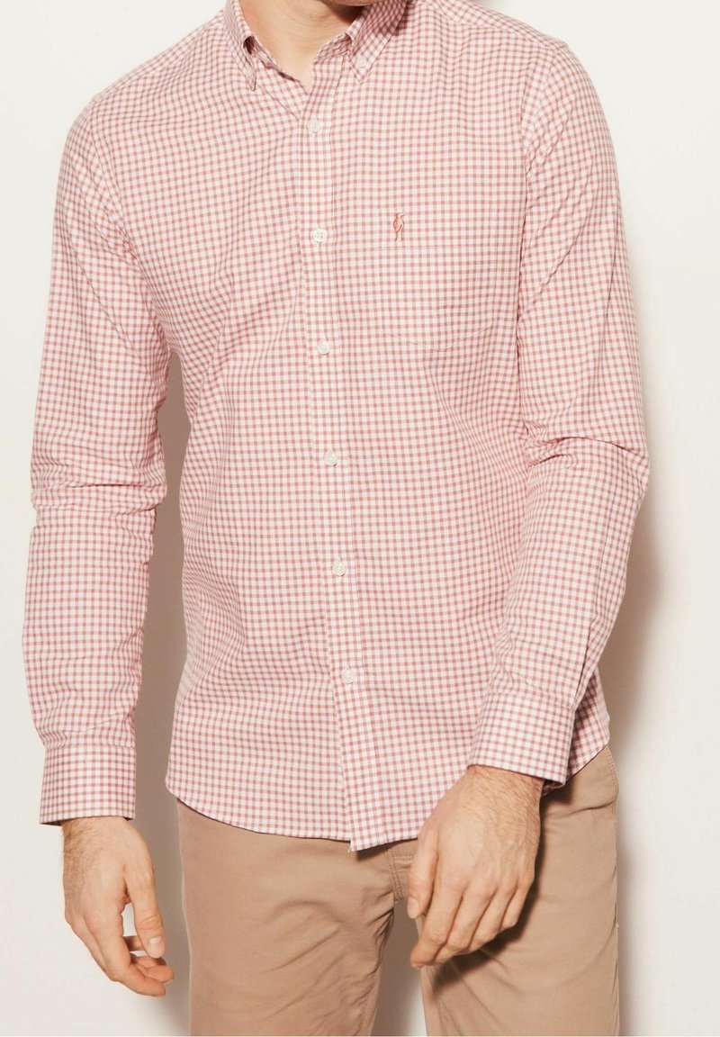 Homme portant une chemise à manches longues à carreaux rouges et blancs avec un pantalon beige, debout devant un fond clair uni.