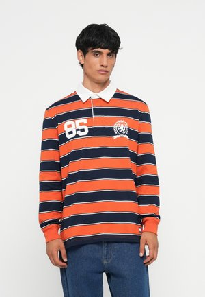 Langermet polo skjorte med oransje og marineblå striper, hvite detaljer, krage og logo. Stylert med blå jeans og klassisk passform.