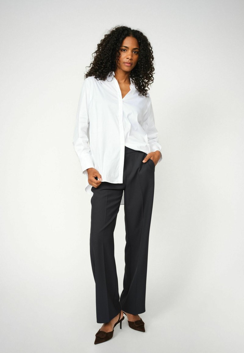 Weißes Hemd mit langen Ärmeln, kontrastierenden dunklen Hosen und braunen hochhackigen Schuhen, vor einem hellgrauen Hintergrund.