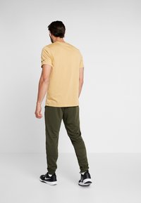 Ljusgul t-shirt med kort ärm i kombination med mörkgröna joggers och svarta sportskor, sedd bakifrån. Enkel, avslappnad klädsel.