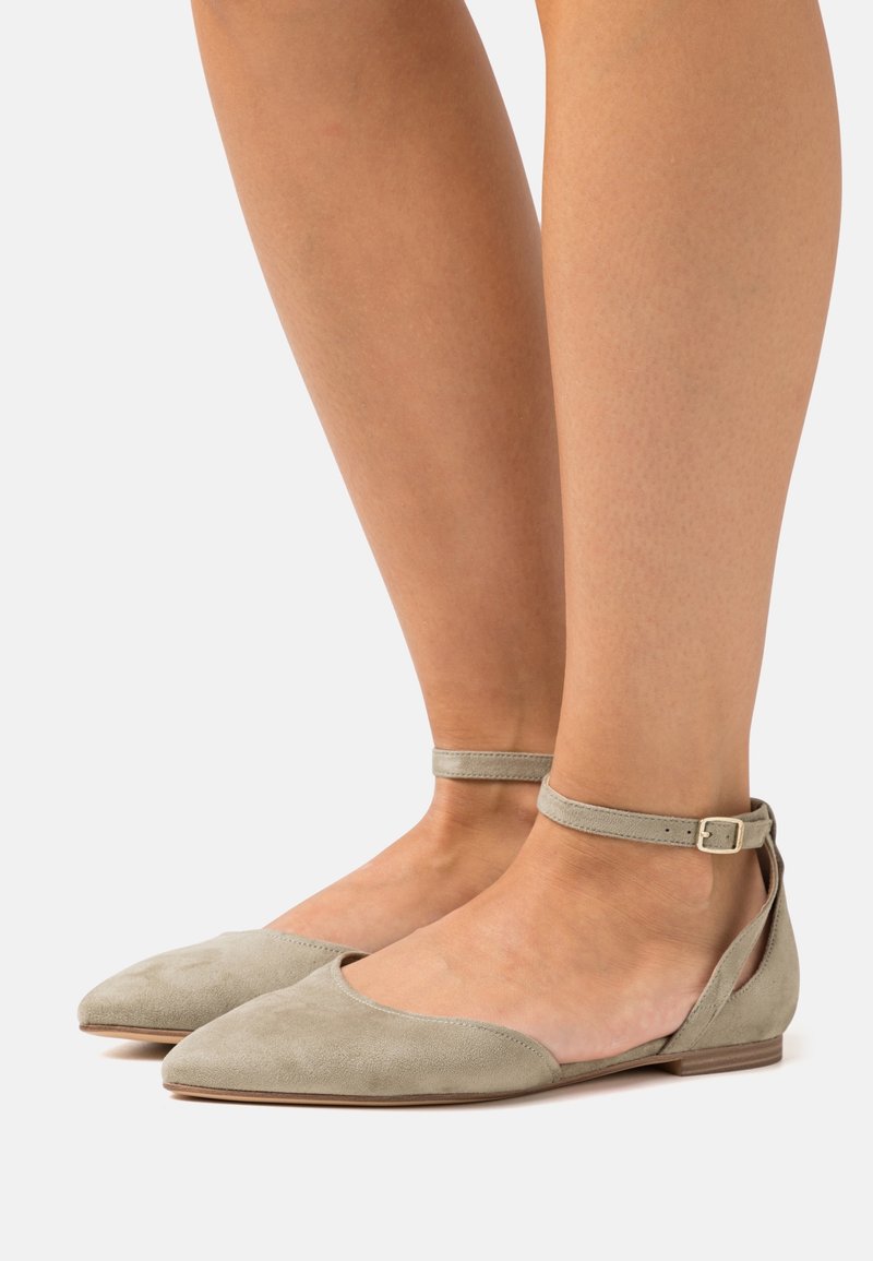 Ballerines beiges en daim avec bouts pointus, brides autour de la cheville et petits talons en bois, portées pieds nus sur un fond blanc.