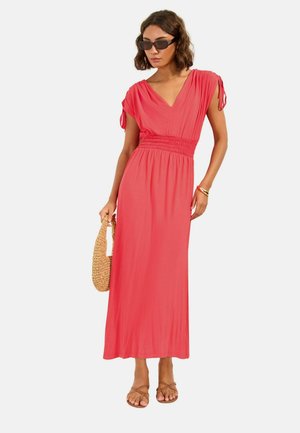 Nő korallrózsaszín maxi ruhában, összegyűrt derékkal és ujjal, fonott kézitáskát tart, szandált és fekete napszemüveget visel.