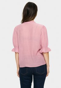 Rosa Baumwollbluse mit Rüsche Ärmeln und gewelltem Spitzenbesatz. Weicher Stoff, leicht und lässiger Schnitt. Getragen mit dunklen Jeans.