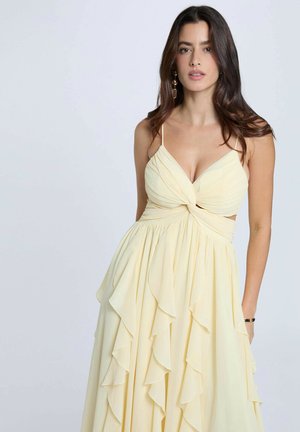 Donna con capelli lunghi e scuri, che indossa un abito giallo pallido senza maniche con gonna arricciata e corpetto annodato, in piedi davanti a uno sfondo chiaro.