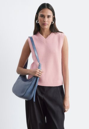 Femme portant un pull sans manches rose et un pantalon noir, tenant un sac à bandoulière bleu sur un fond blanc uni.