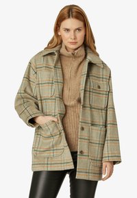 Giacca a quadri verde e beige con dettagli arancioni, caratterizzata da un design con colletto, tasche frontali e una vestibilità comoda sopra un maglione beige.