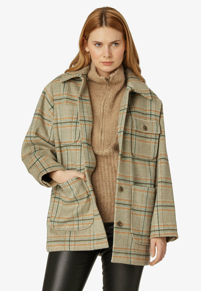 Giacca a quadri verde e beige con dettagli arancioni, caratterizzata da un design con colletto, tasche frontali e una vestibilità comoda sopra un maglione beige.