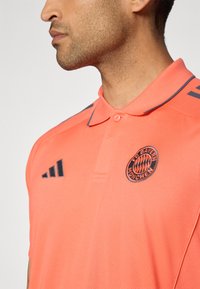 Polo rose en tissu texturé, avec un col marine, le logo du FC Bayern München et trois rayures marine sur l'épaule.