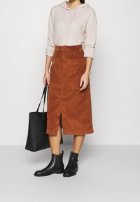 Falda midi de pana marrón con abertura frontal y dos bolsillos, combinada con un hoodie de canalé gris claro y botas negras hasta el tobillo. Se muestra un bolso tote negro.