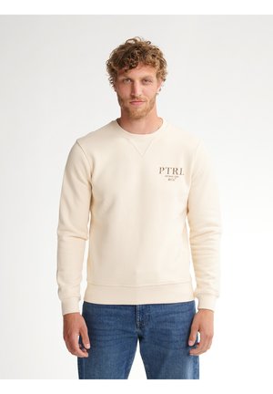 Man met krullend haar die een crèmekleurige sweater met PTRl-logo en een blauwe spijkerbroek draagt, staand tegen een effen witte achtergrond.