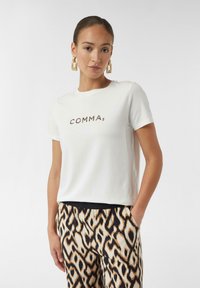 Weißes T-Shirt mit kurzen Ärmeln, bedruckt mit "COMMA" in strukturierten schwarzen und braunen Buchstaben. Kombiniert mit gemusterten, lockeren, mehrfarbigen Hosen.
