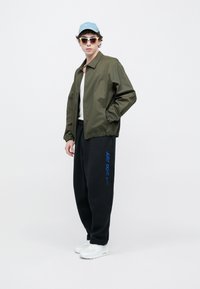 Olivzaļš bumbvedēja stila džekets, balta T-krekls, melnas sporta bikses ar zilām "JUST DO IT" burtiem, balti sporta apavi, gaiši zila cepure un saulesbrilles.