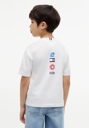 Balra néző fiú, fehér rövid ujjú ingben, melyen kék hullámok, zászlók, nap ikonok és a "EST.85" szöveg található a háton, világoskék farmerral párosítva.