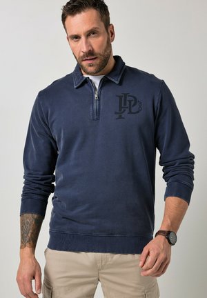 Mand iført marineblå kvart-lynlåstrøje med logo, beige cargobukser, ur og en tatovering på hans venstre underarm mod ensfarvet baggrund.