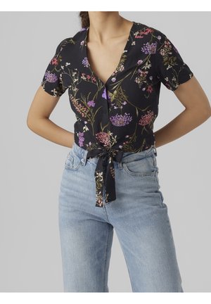 Kvinde iført en sort blomsterprintet kortærmet skjorte knappet op og bundet i taljen, parret med lyseblå højtaljede jeans.